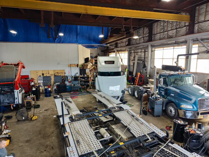 Busy shop floor with a blue tractor, empty car-hauler trailer, and equipment in for service.