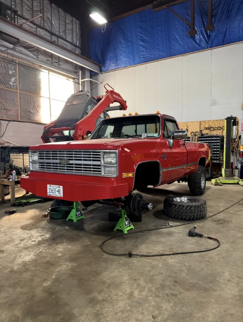 Red Chevrolet pickup on jack stands in the Top Guns bay, front wheel off and a tire on the floor.