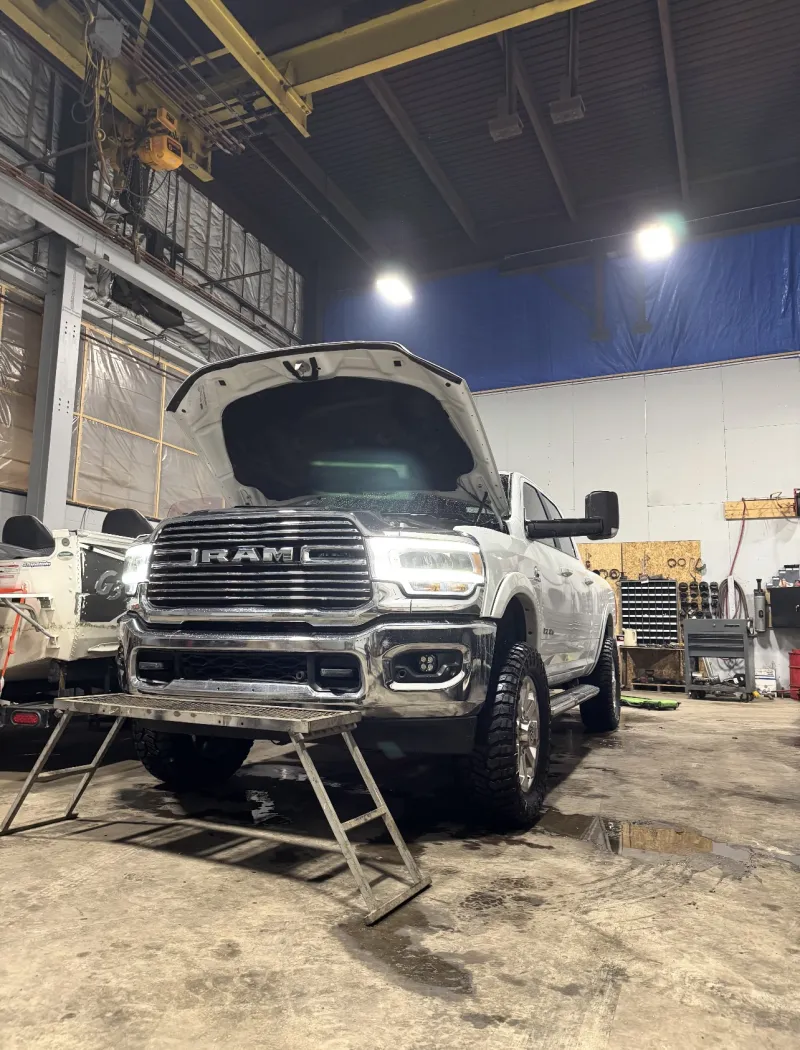 White RAM in the shop with the hood up and headlights on.