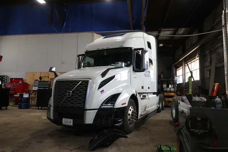White Volvo with the driver-side bumper off and the part on the shop floor.